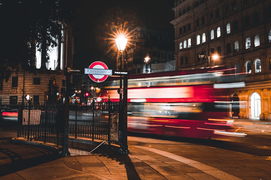 A nighttime street scene near Earls Court Station featuring a classic London underground sign mounted on a black pole, illuminated by warm street lamps. In the background, historic buildings with lit 