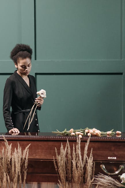 A young woman with dark curly hair, dressed in a black coat, holds a small bouquet of white flowers with green stems, standing in front of a dark wooden table with a floral arrangement laid out on it.
