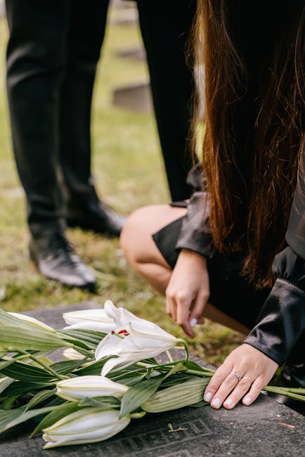 A person with long brown hair, dressed in black, is kneeling on the ground, carefully placing a floral arrangement consisting of white lilies and green leaves onto a flat surface, likely at a funeral 