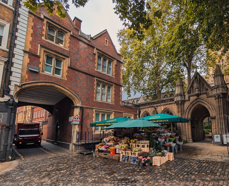 A street scene outside an ornate, historic building with a large arched gateway, featuring a flower stall set up beneath a green umbrella. The stall displays an array of fresh, vibrant flowers includi