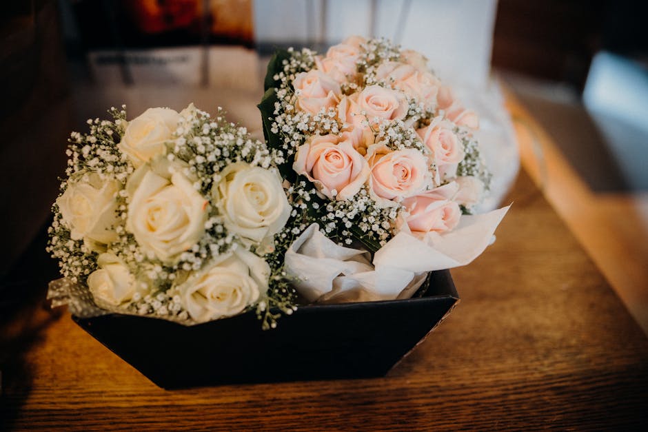 A floral arrangement featuring two bouquets of fresh roses, one with creamy white roses and the other with soft pink roses, each surrounded by delicate sprigs of baby's breath. The bouquets are arrang