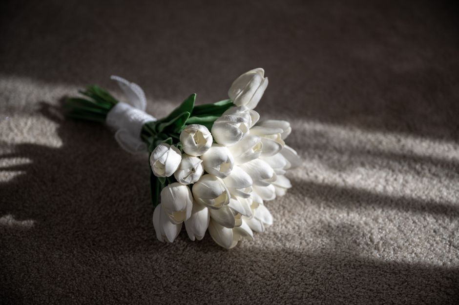 A bouquet of fresh white tulips with closed petals, bound together with white ribbon and green stems, rests on a beige textured carpet. The flowers are arranged with some tulips slightly leaning, show